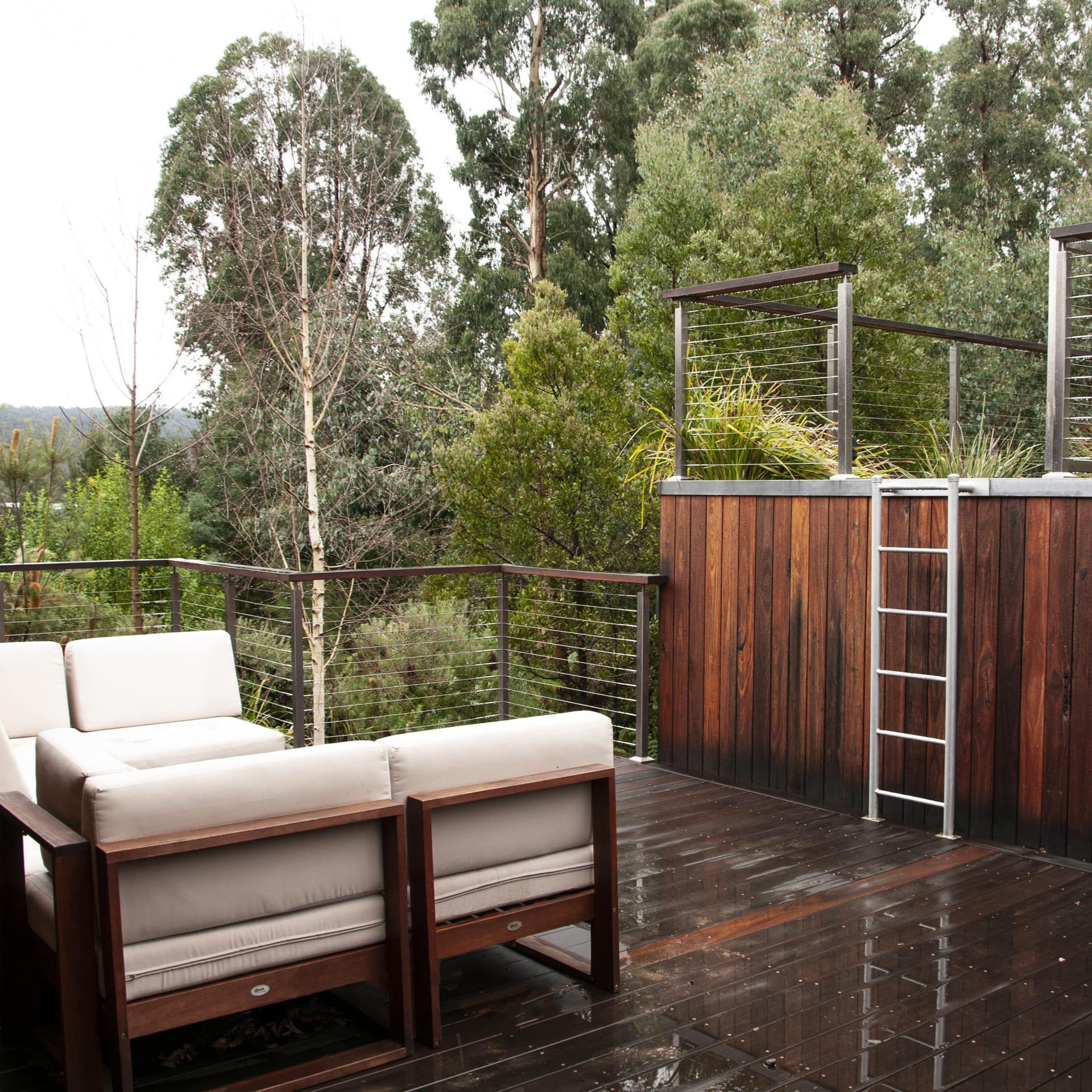 Wood patio space with white canvas sectional, a roof garden with metal ladder for access, surrounded by thick forest.