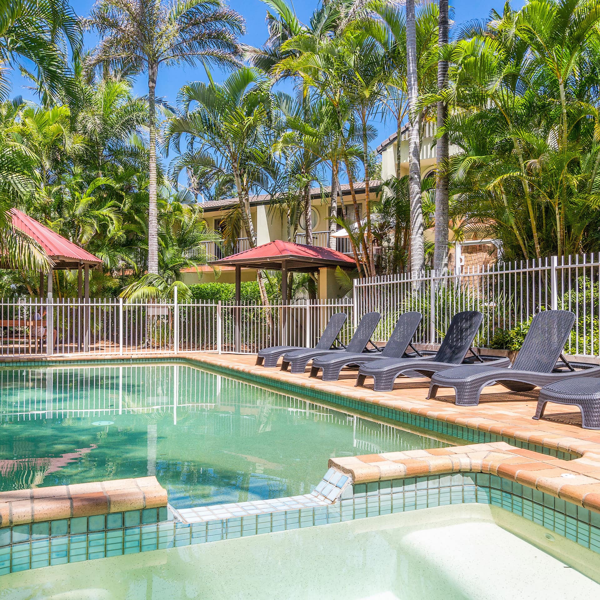 Sunny view of pool and patio space, tan tile, black lounge chairs, enclosed by white iron fence and palm trees.