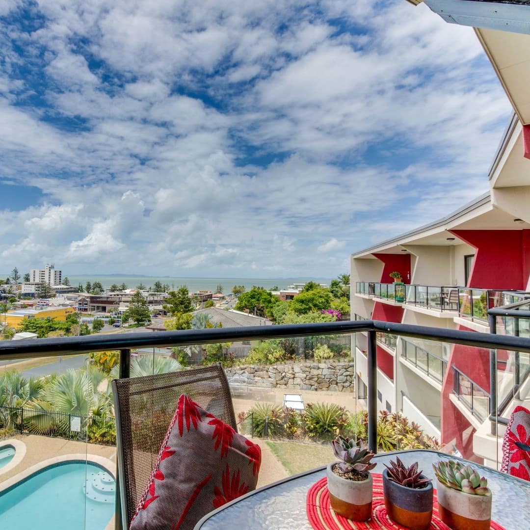 View of pool, neighborhood, and ocean from apartment balcony with glass railings and dining set.