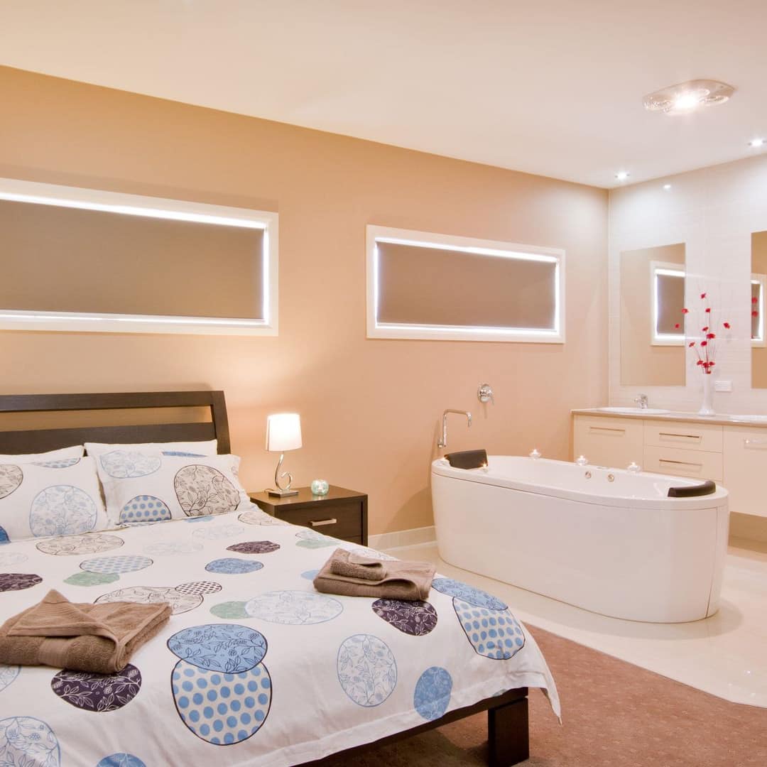 Bright bedroom with tan walls and carpet, a queen size bed, blue and white bedding, a tub, and his and hers bathroom vanity.