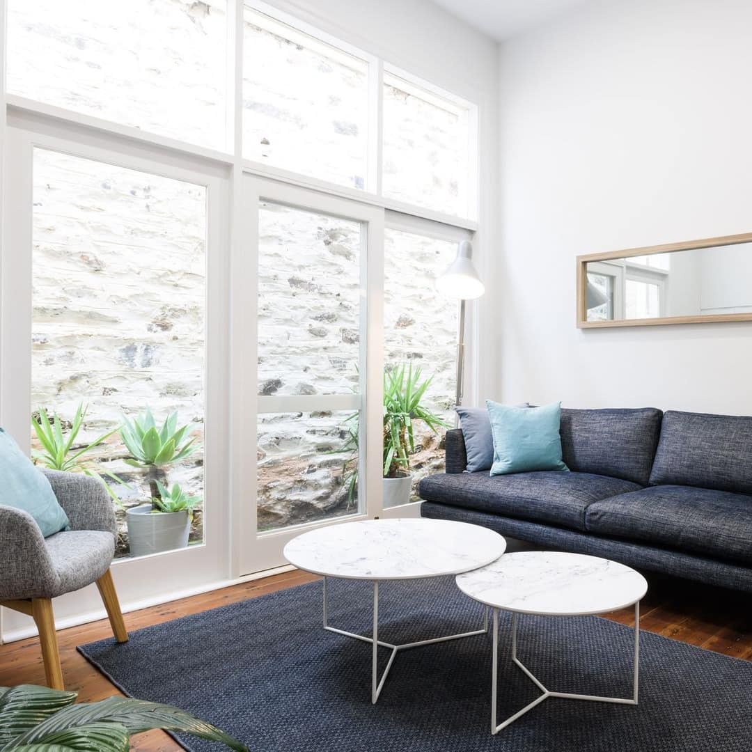 Open living room with hardwood floors, white walls, grey sofa, marble-topped table, and tall windows.