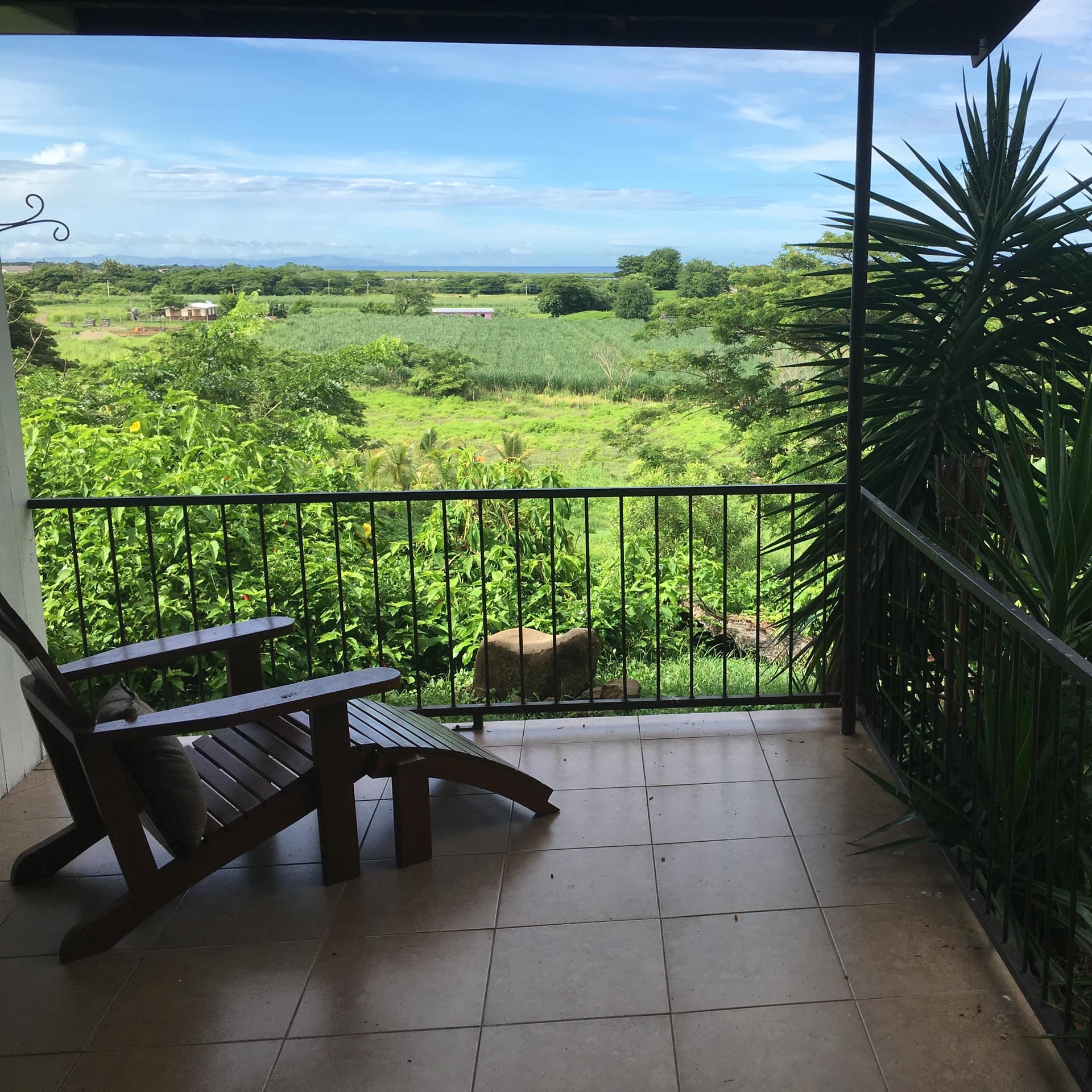 A covered deck with beige tile and wooden lounge chair, overlooking a lush rural landscape.