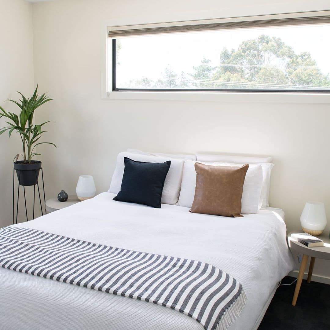 Bedroom with black carpet, a queen-size bed, white bedding and striped blanket, a potted plant and wide windows.