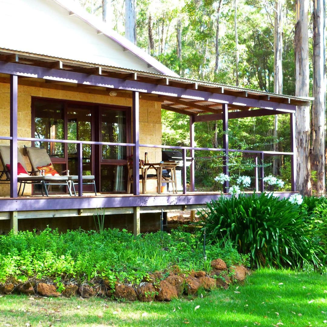 Exterior of tan cottage with covered wrap-around porch, beige lounge chairs, and a landscaped yard with lush vegetation.