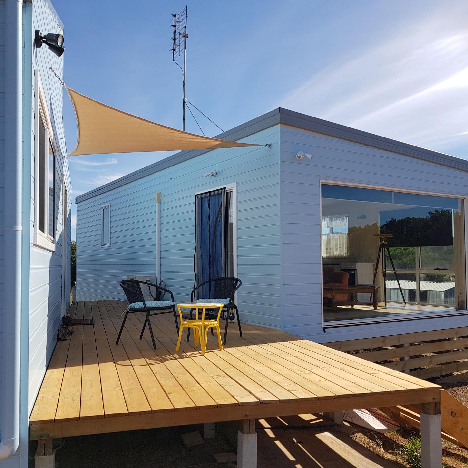 Two modern, blue cottages connected by a wooden deck with black chairs and a yellow table, and a canvas covering.