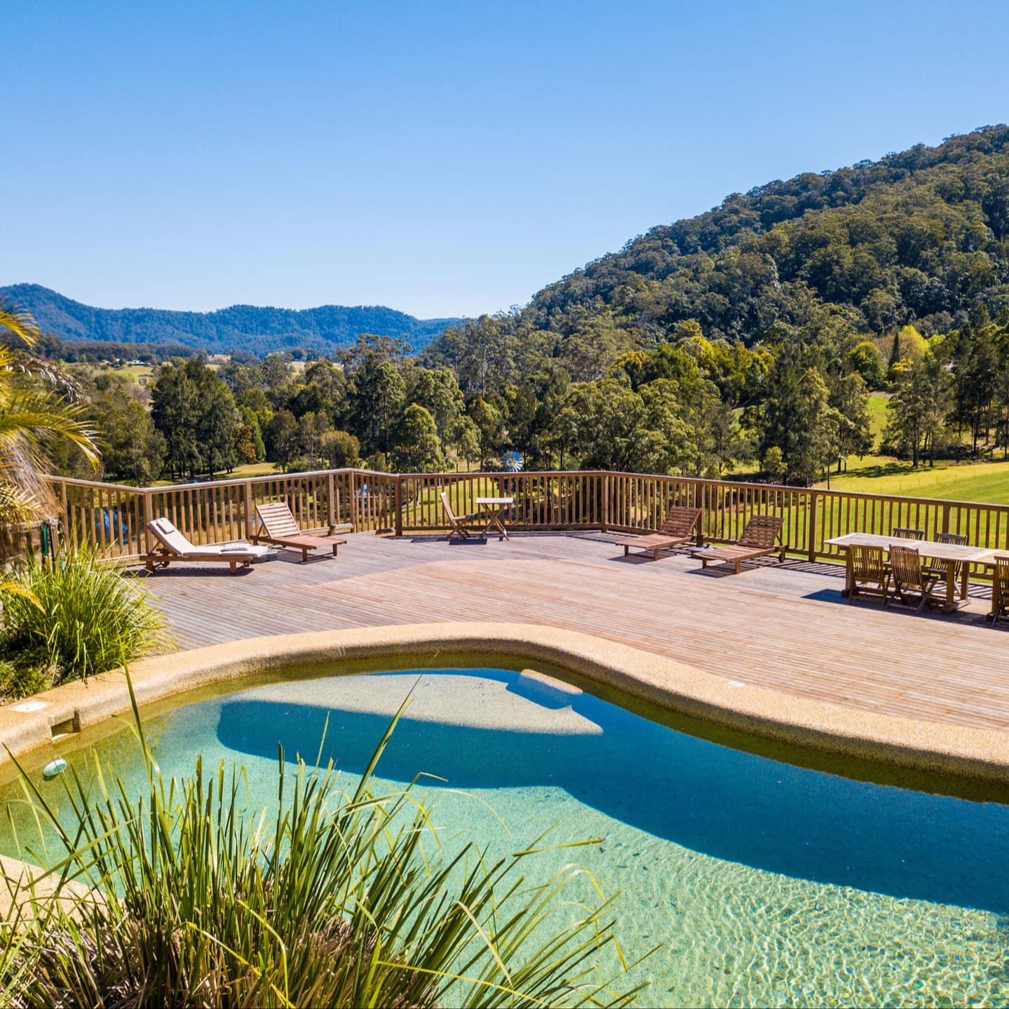Sunny view of the private pool with wood deck, lush tropical vegetation, wood lounge chairs and dining set, and forest views.