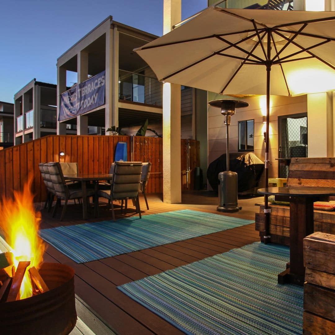 Back deck of 2-storey house with rustic wood seats, bright pillows, an umbrella, outdoor dining table and a burning fire.