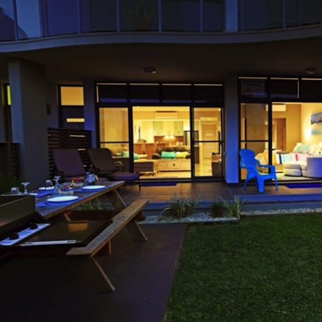 Grassy backyard of apartment with covered patio, outdoor dining, grill, lounge chairs, large windows and a sunset.