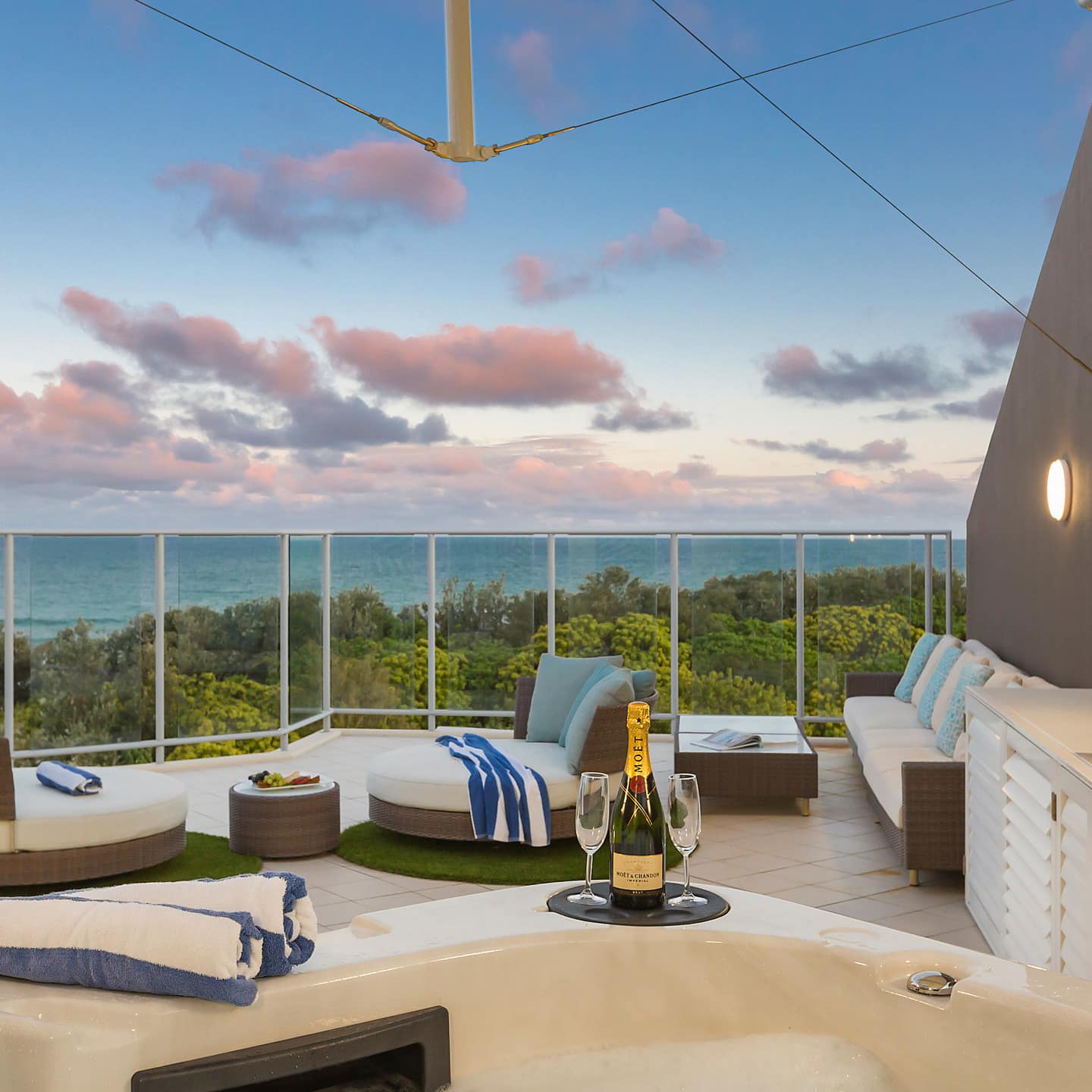 A balcony with glass railings, beige tile, modern wicker furniture, hot tub and views of a sunset setting over the ocean.
