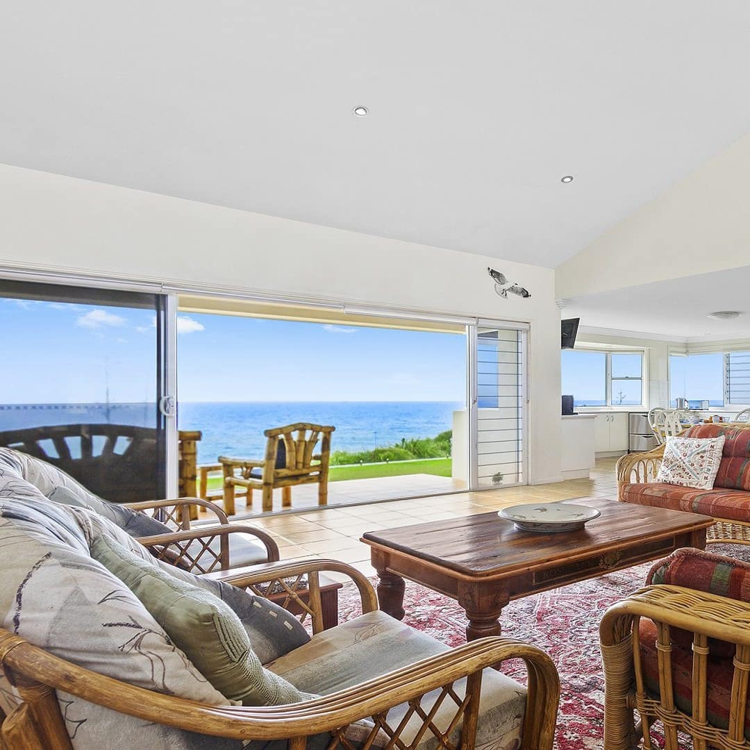 Open living and dining room with rattan sofas and chairs, Persian rug, dining hutch, and sliding glass doors to waterfront.