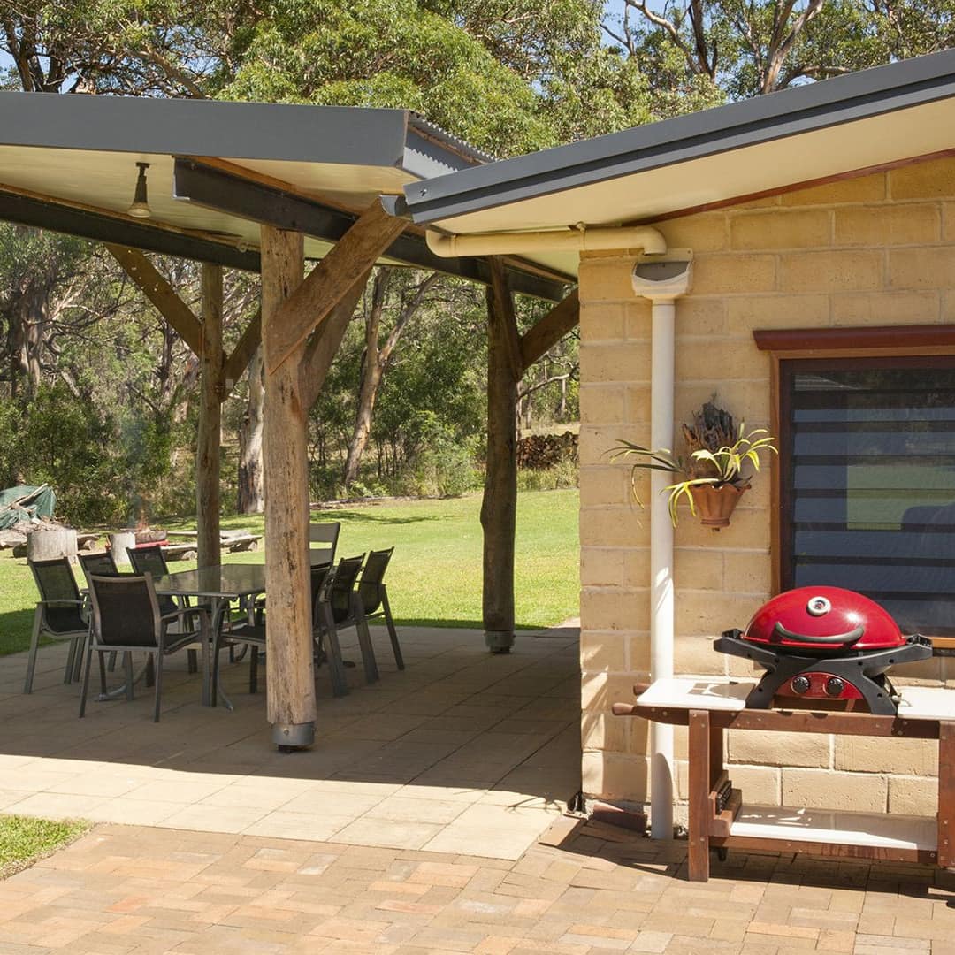 View of exterior of home, covered patio with beige tile, glass dining set, surrounded by lush lawn and trees.