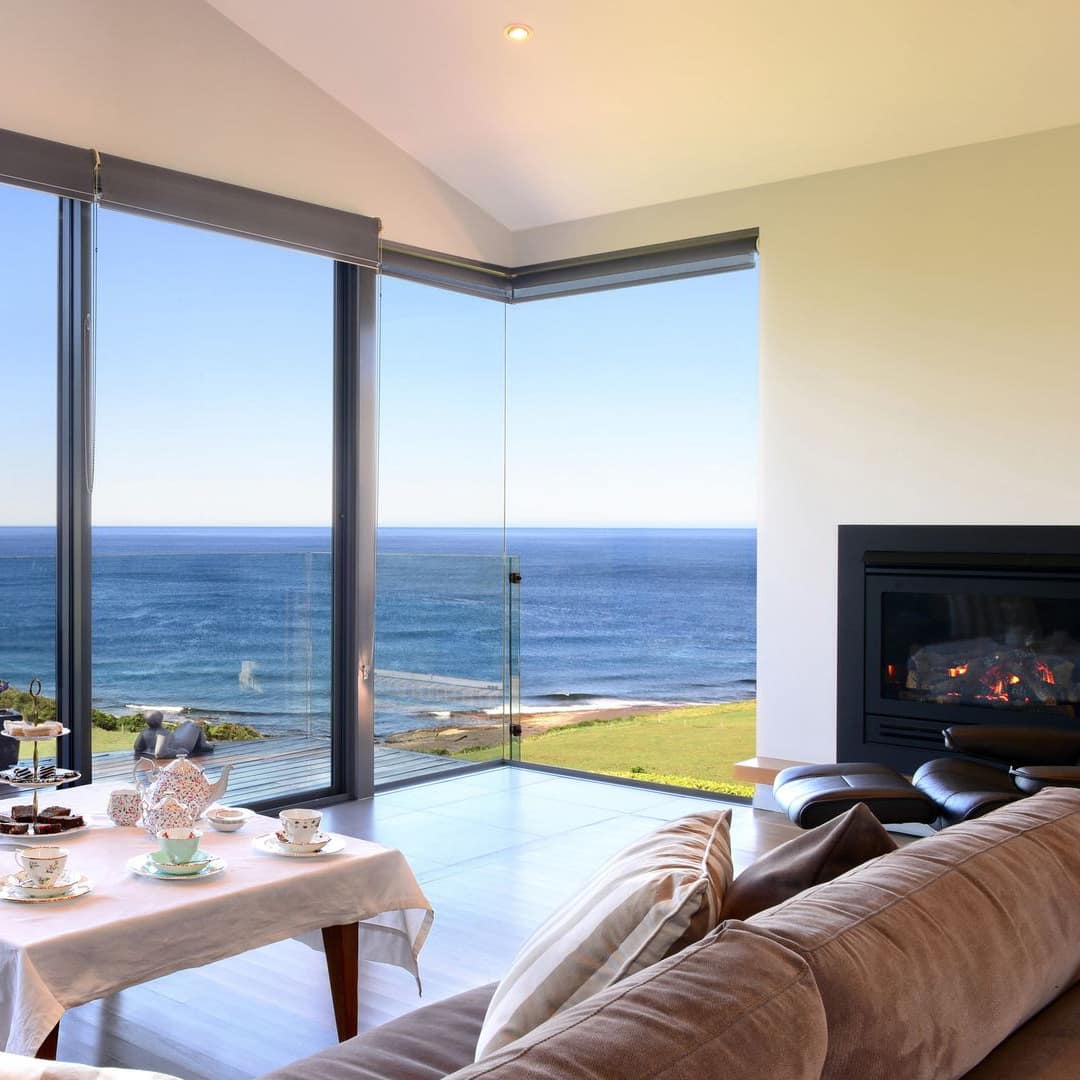 Large windows with ocean views, brown L-shaped sectional, coffee table set with high tea, electric fireplace.