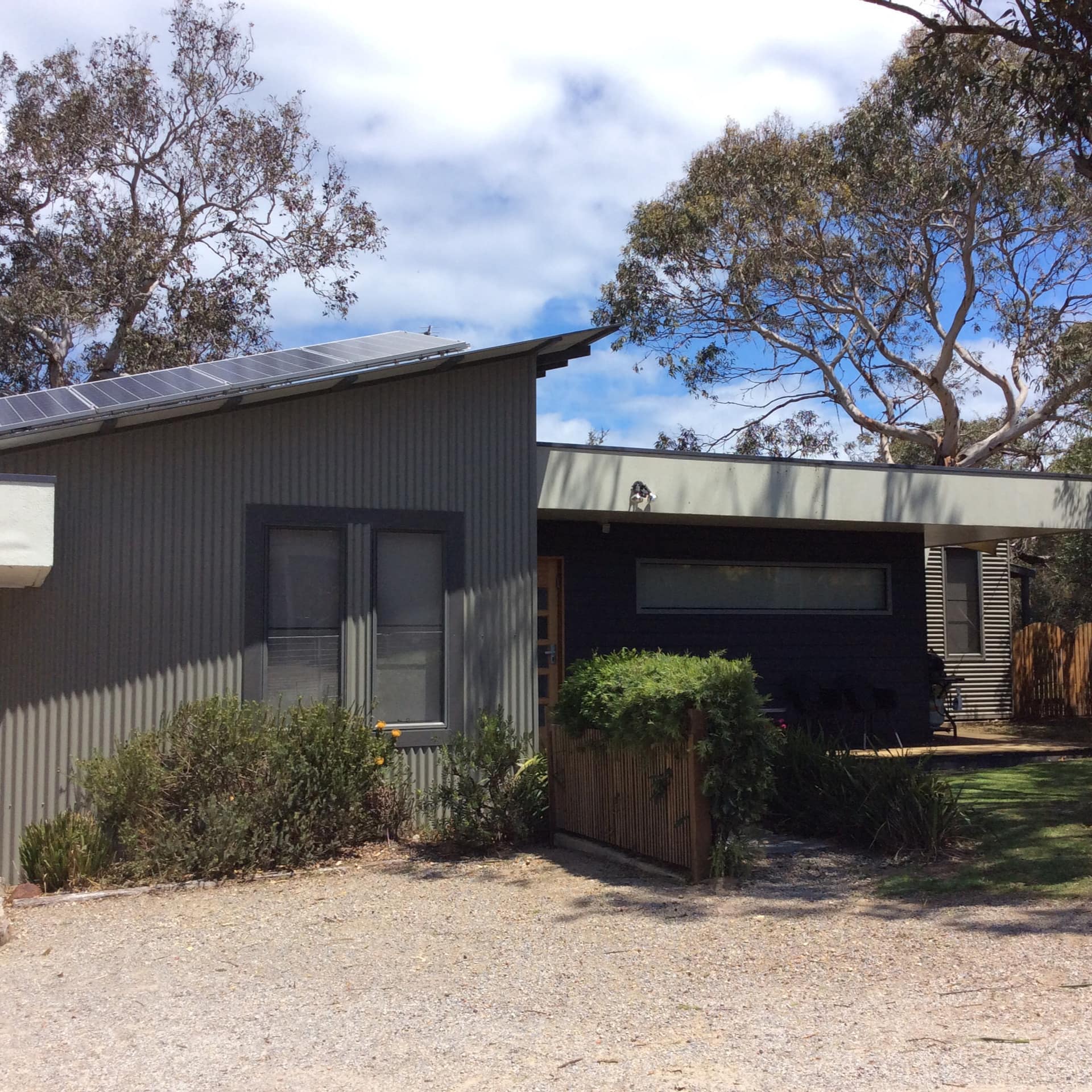 Modern home with grey siding, solar panels on the roof, gravel driveway, surrounded by trees.