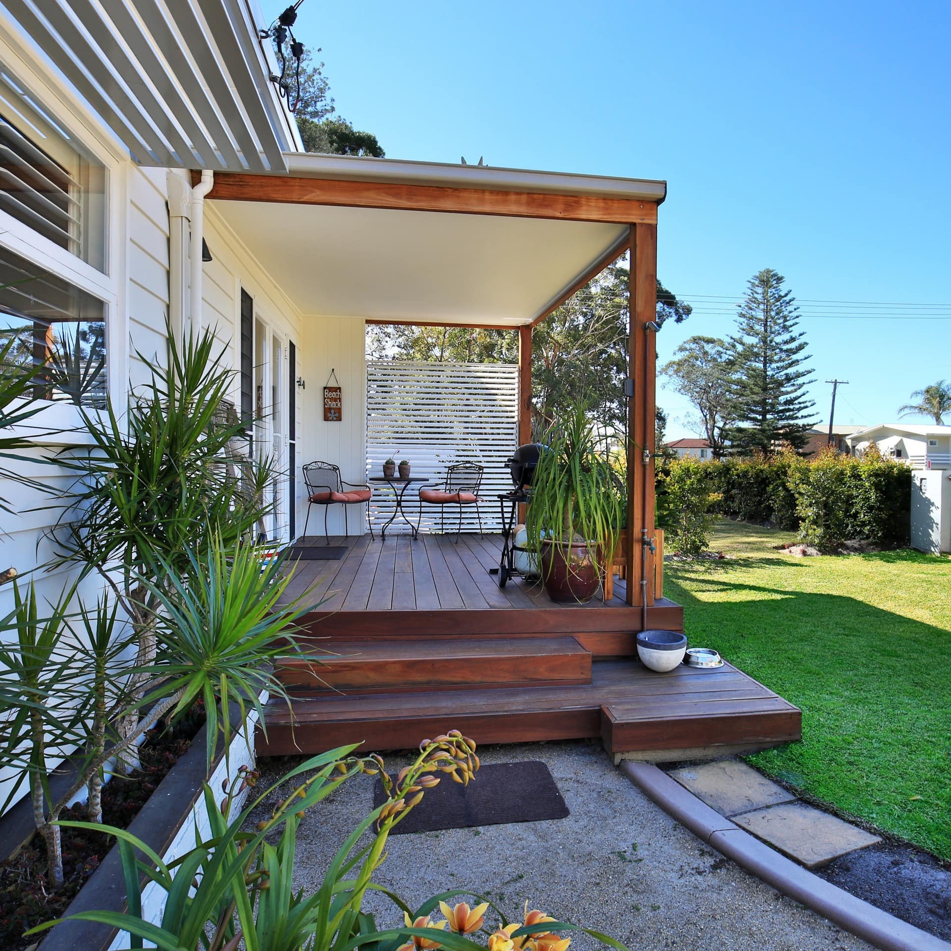 Covered, hardwood deck in a landscaped backyard, pebble gravel path, potted tropical plants, and manicured shrubbery.
