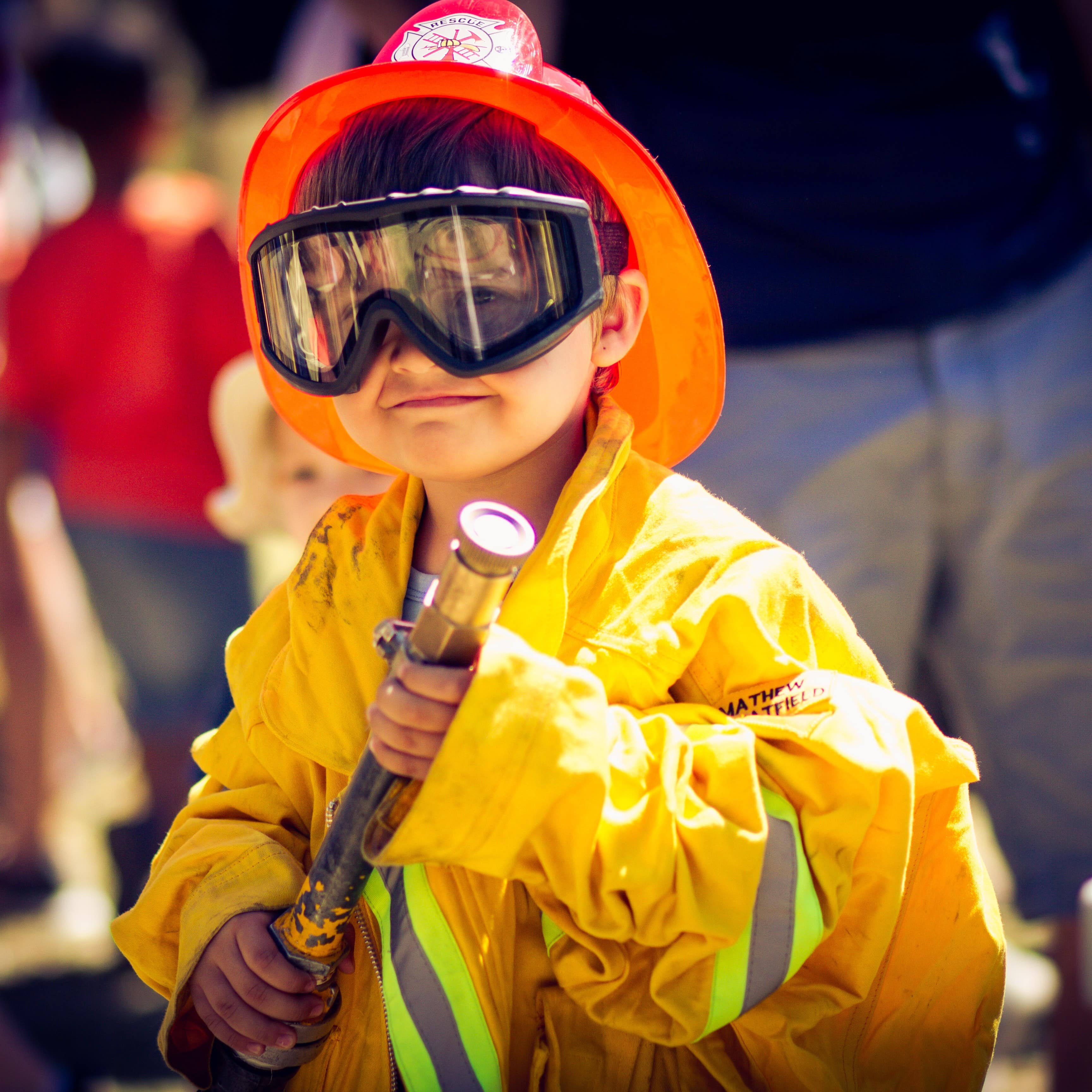 Young boy dressed in yellow fire gear with goggles and helmet holding a fire hose