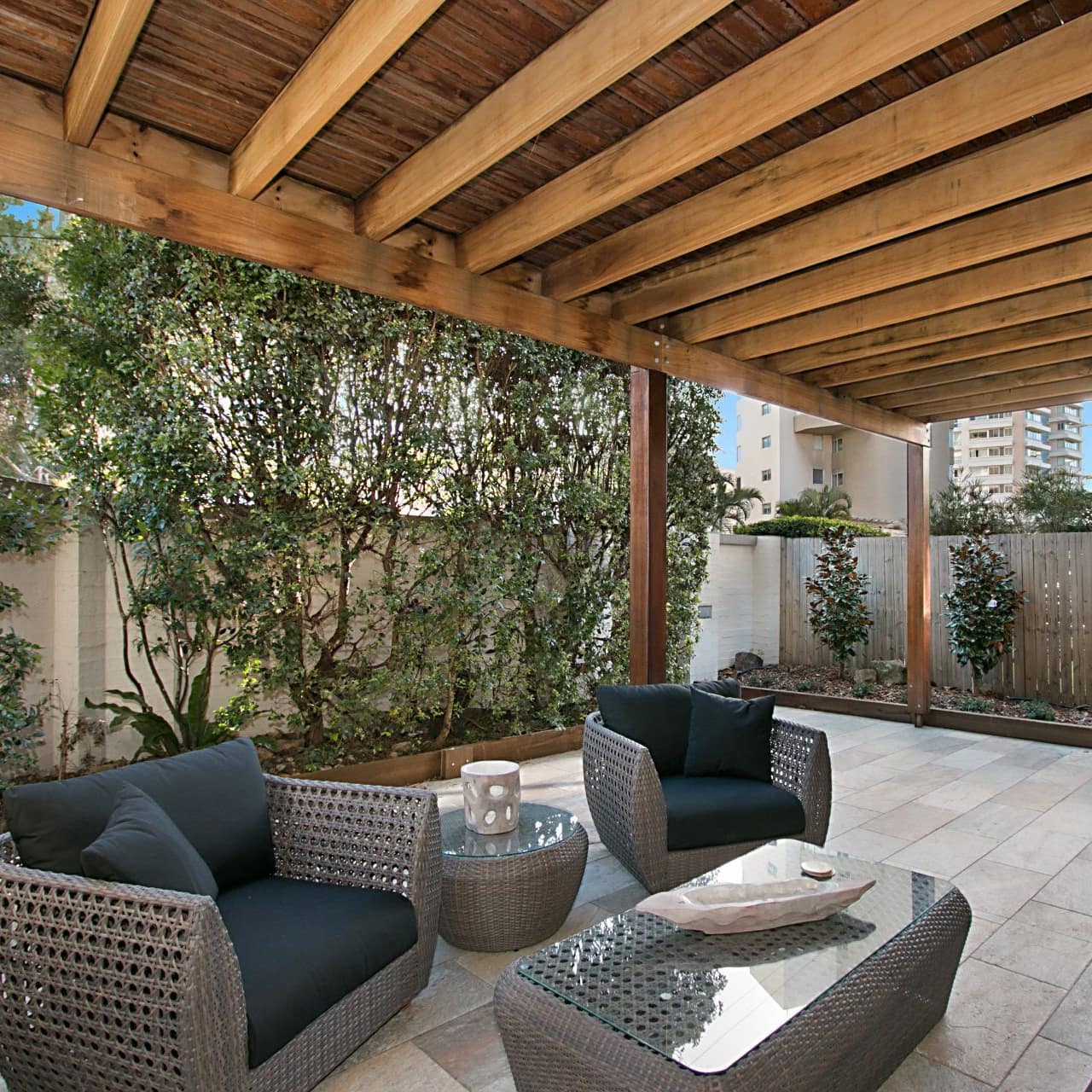 A covered patio with grey tile floor, matching grey wicker chairs, sofa, and tables, and plant beds with small trees.