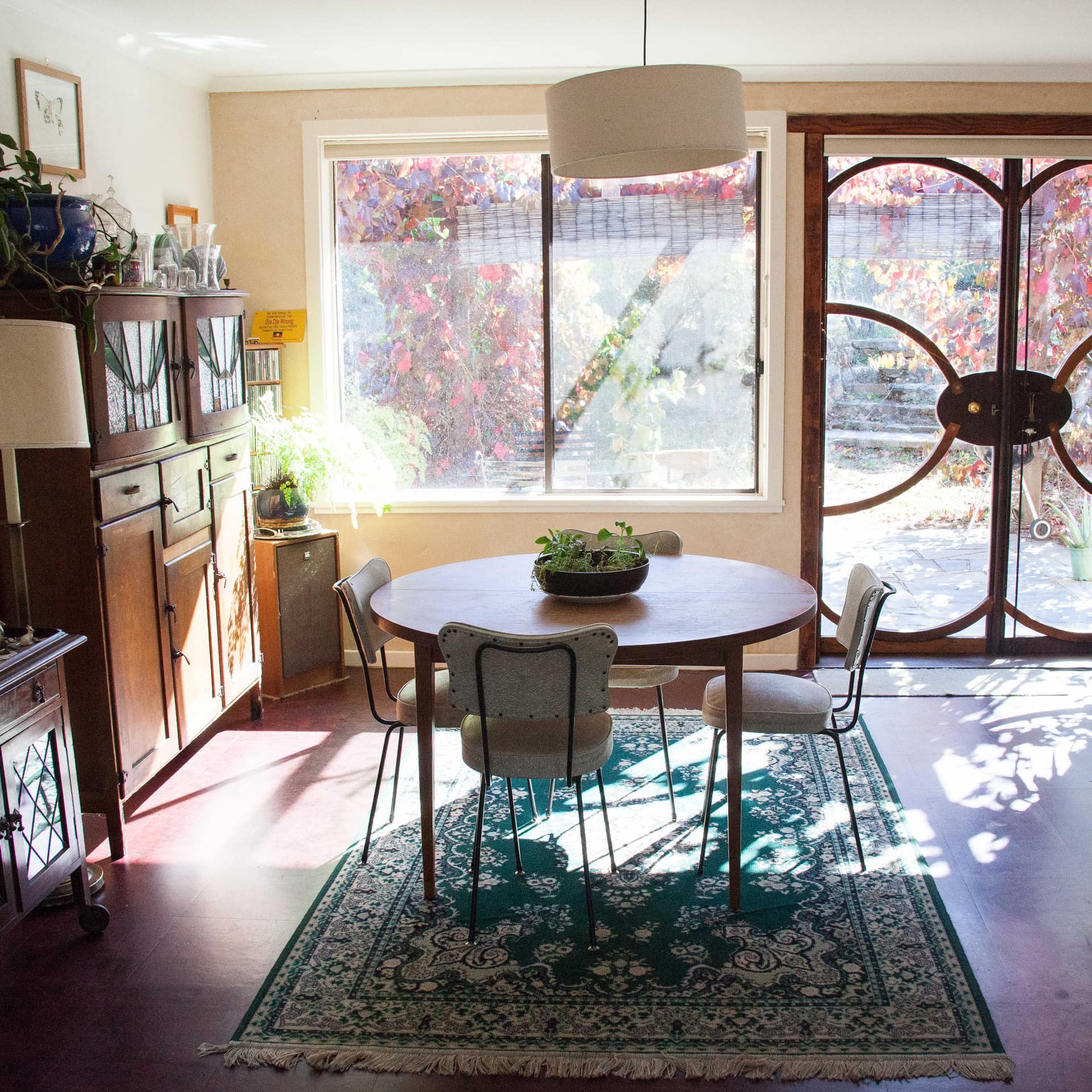 Open dining room with round dining table, antique wooden cabinet, green persian rug, and double glass door to patio.