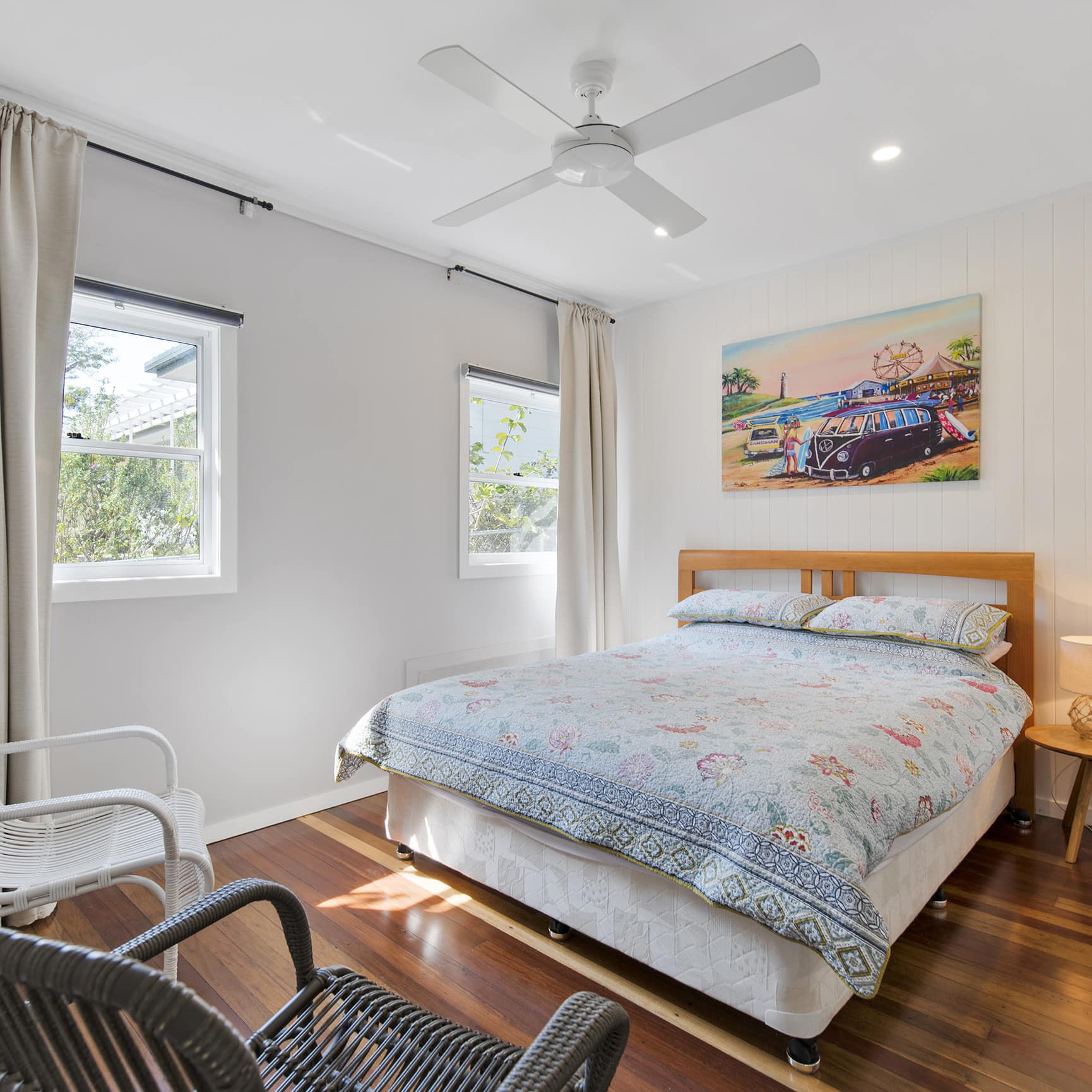 Sunny bedroom with hardwood floors, white walls, a queen size bed, light blue bedding, windows, beige curtains.