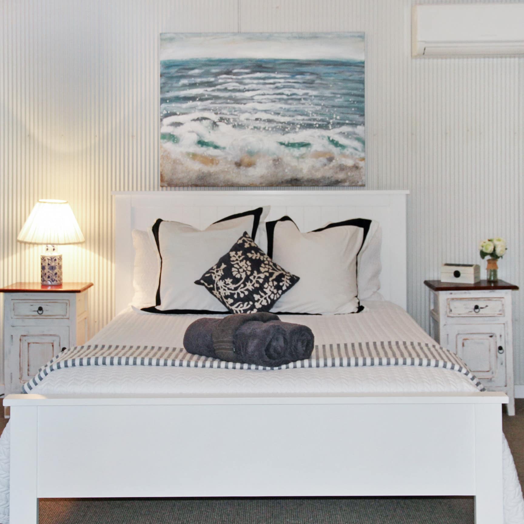 Bedroom with white walls, tan carpet, queen size bed with white linen and black pillows, and a painting of the ocean.