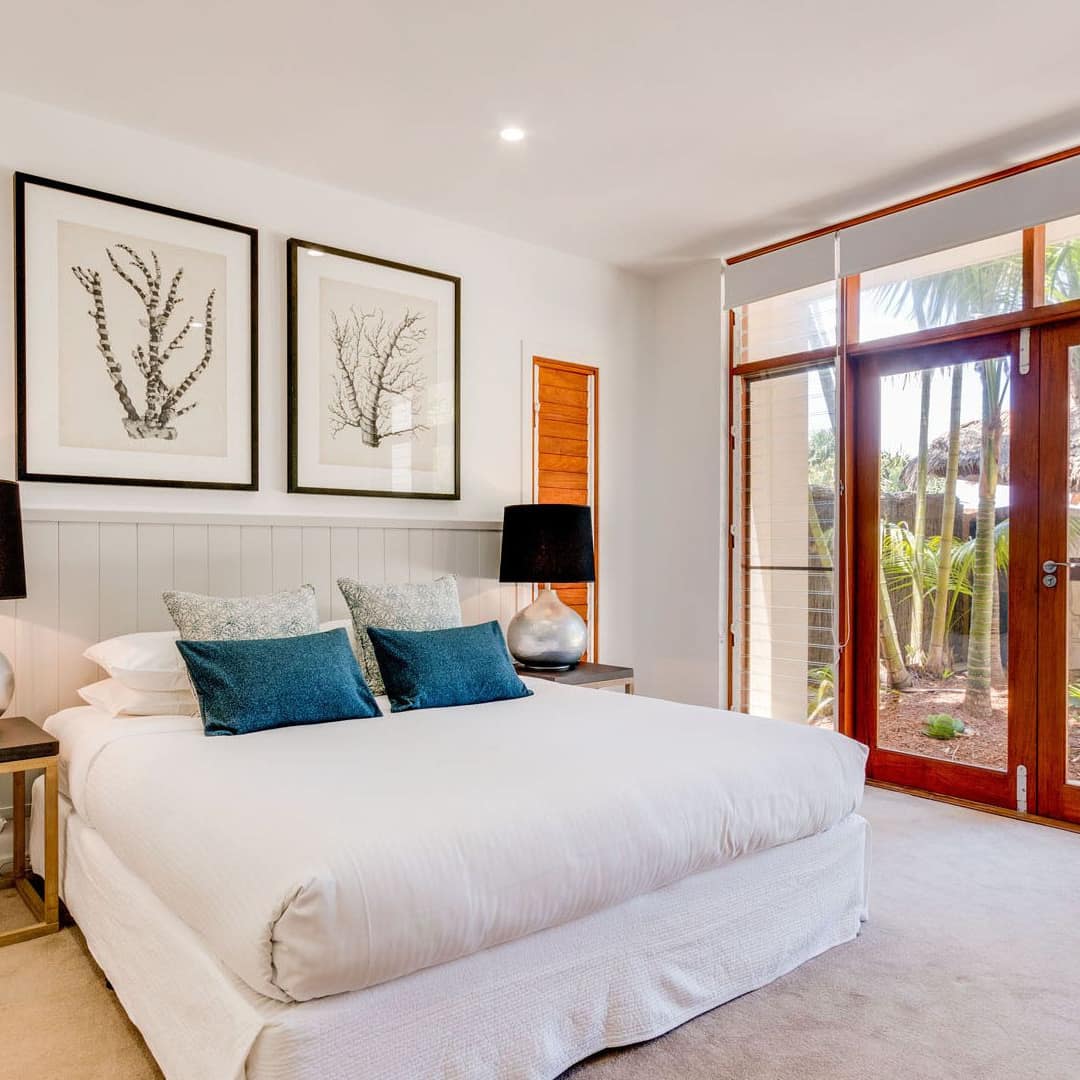 Spacious bedroom with a king size bed, beige bedding and blue pillows, modern decor, and glass doors lead to patio space.