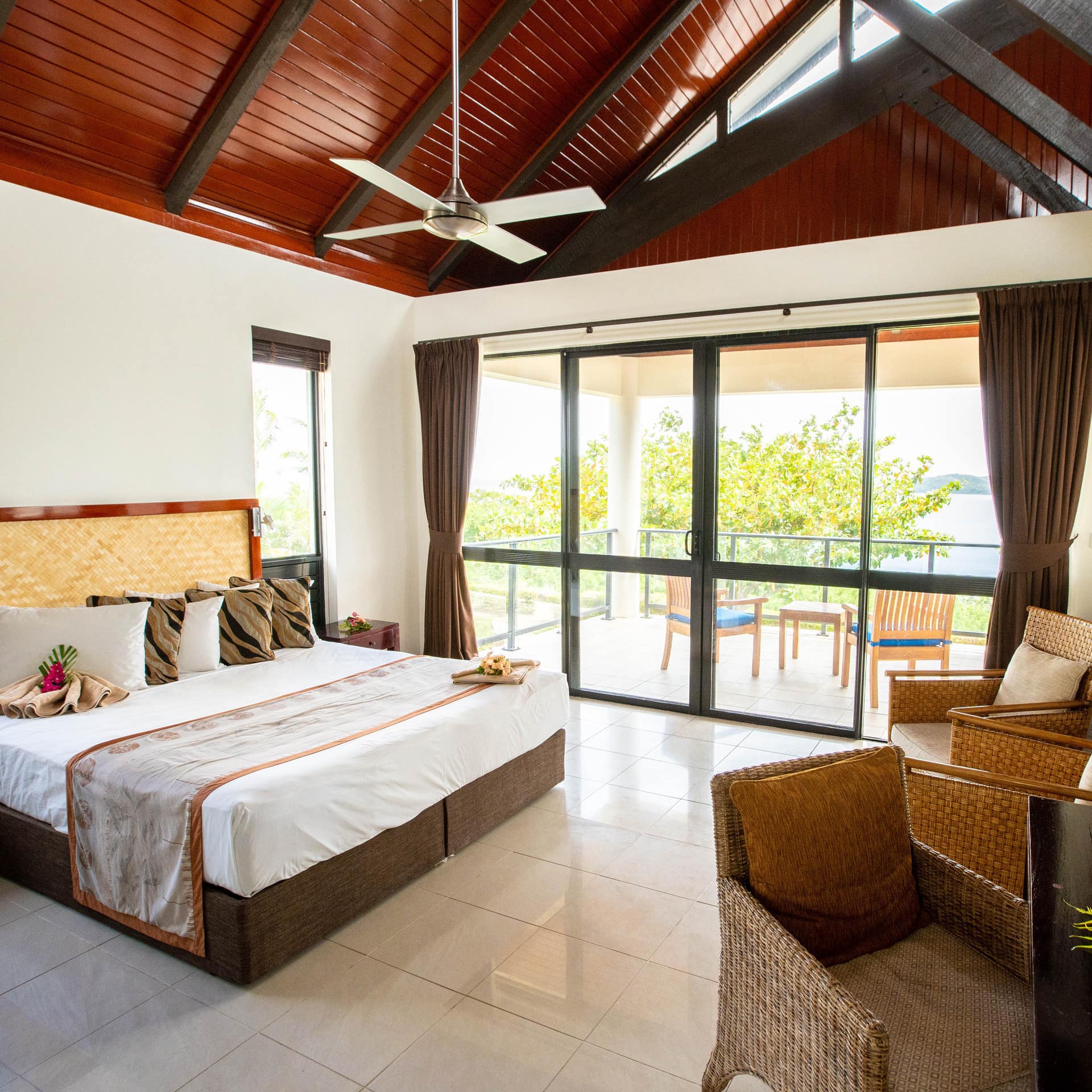 Sunny bedroom with white tile floors, white walls, king size bed, white bedding, wicker furniture, glass door to a balcony.