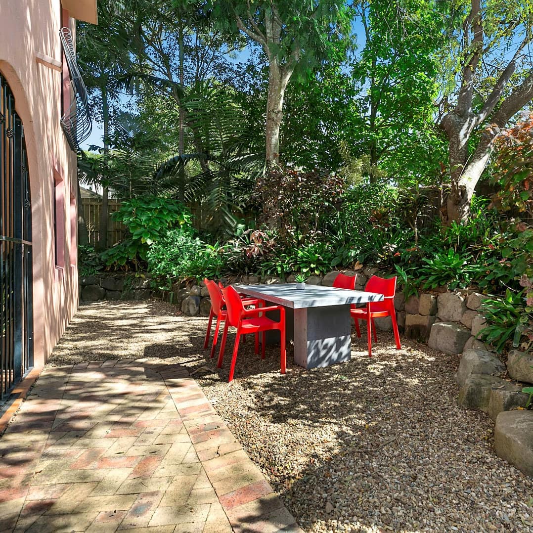 View of a gravel patio space with a grey dining table and red chairs, surrounded by lush greenery.