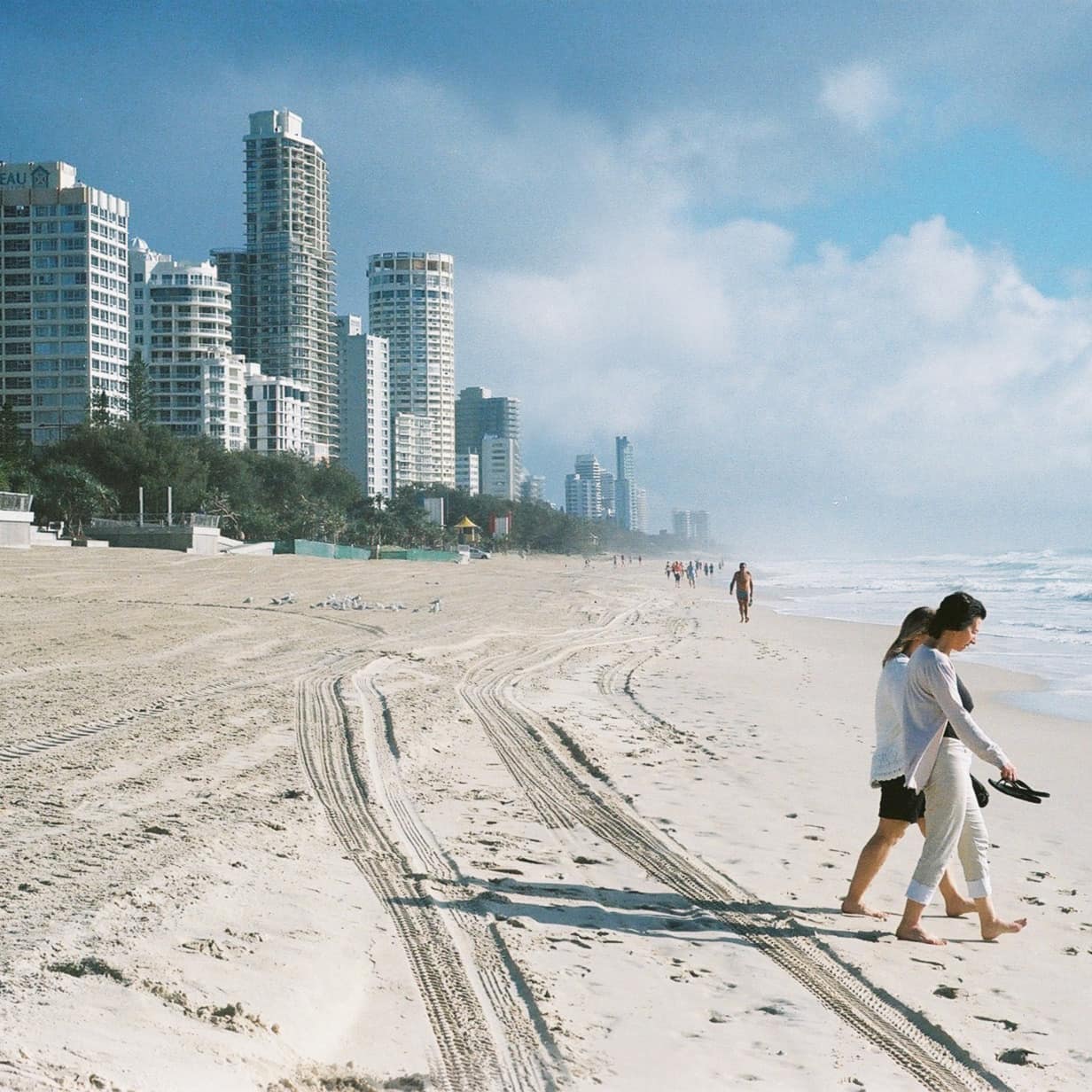 Two women walk on a sandy, white beach with high-rise apartments in the background and waves crashing on the shore