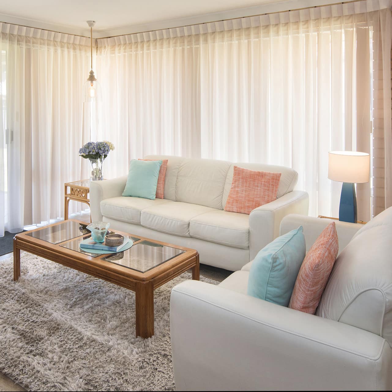 Living room with hardwood floors, floor to ceiling windows with sheer curtains, white sofa and chair, wooden coffee table.