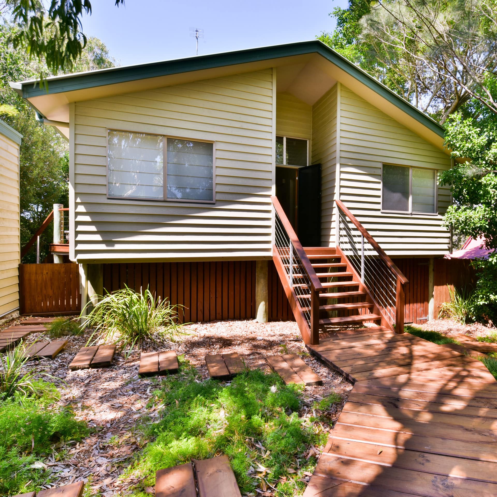 Sunny view of yellow cottage with wooden pathway surrounded by lush greenery.