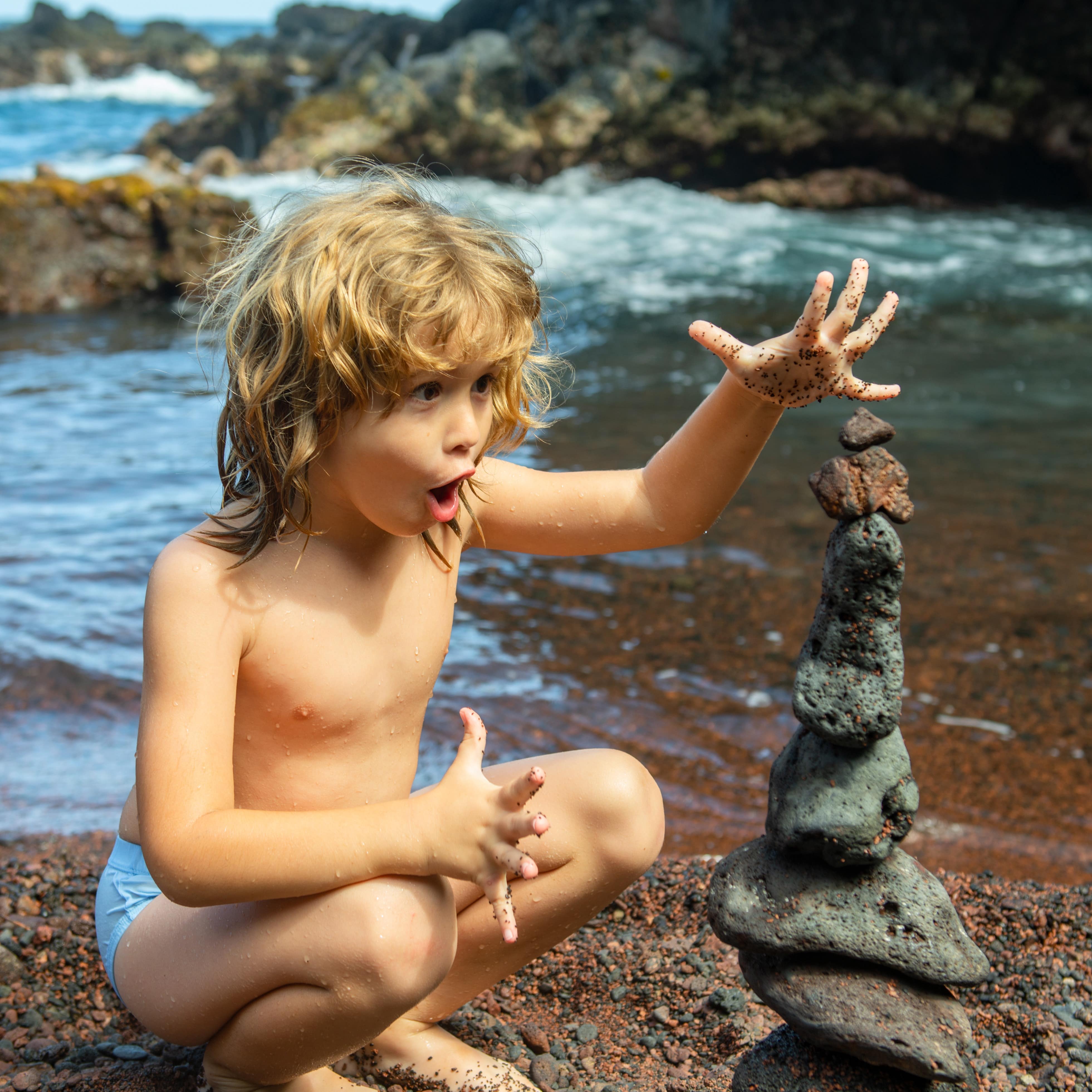 Young boy makes funny face stacking stone pebbles against sea background