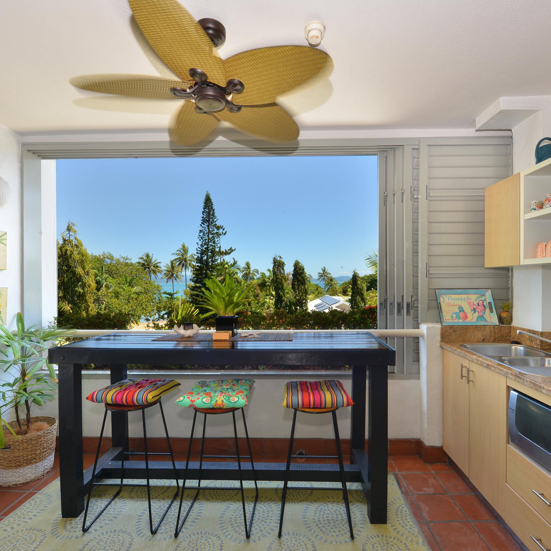 Colorful kitchen, wood cabinets, granite countertops, black bar, stools, wide window with ocean views, bamboo ceiling fan.