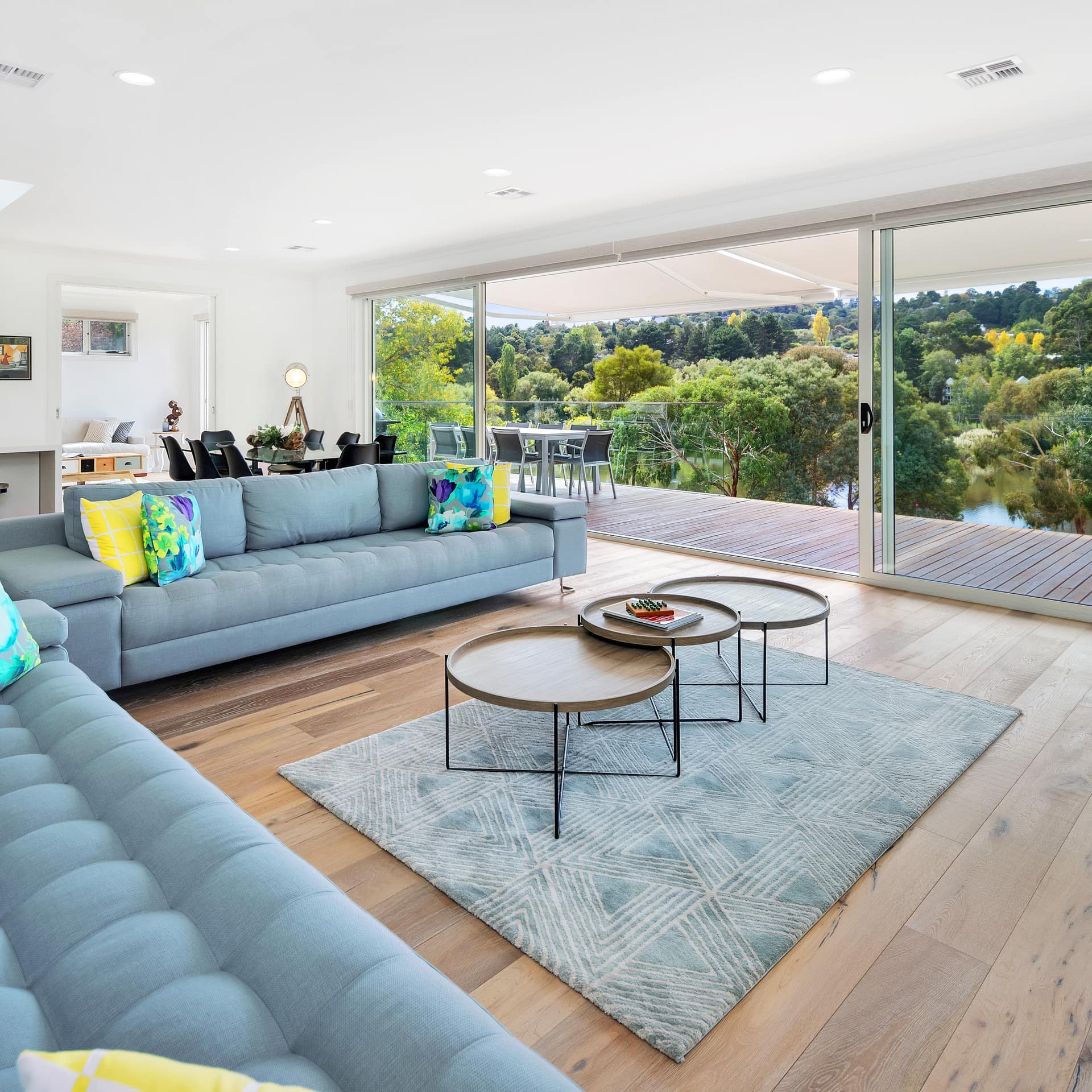Modern, open living room and kitchen with floor to ceiling windows with lake views, hardwood floors, and light blue sofas.