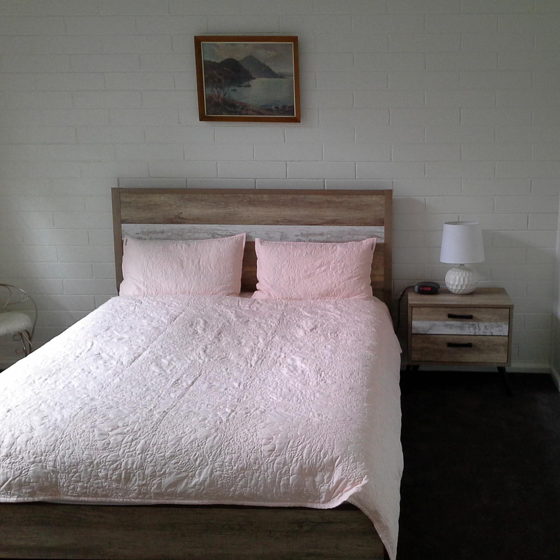 Bedroom with white brick wall, wood-framed queen size bed, pink bedding, wooden bedside table, nautical art.