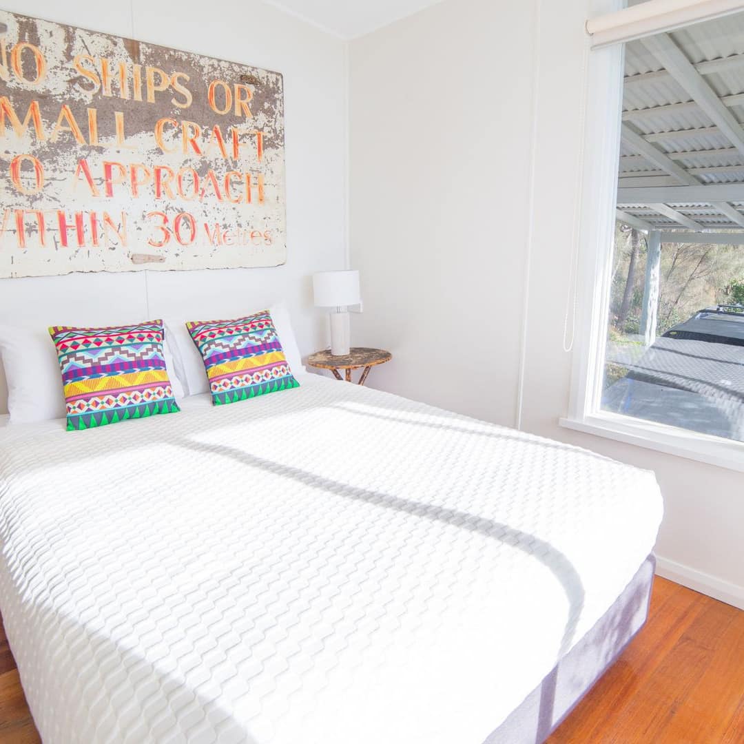 Sunny bedroom with hardwood floors, white walls, a king-size bed with white bedding, colourful pillows, vintage art.
