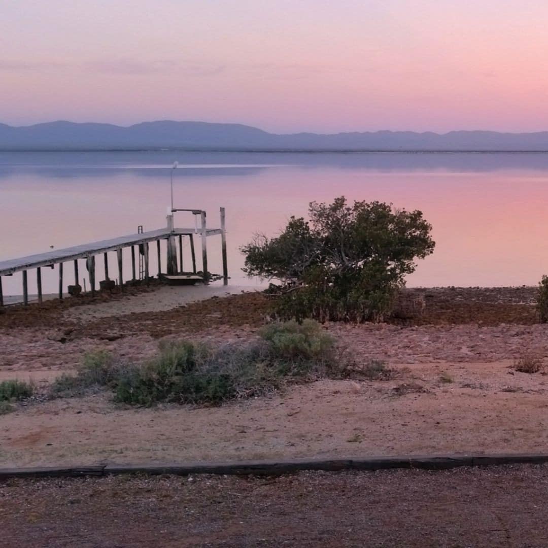 The view from the front porch of a pink sunset reflecting on the ocean, a mountain range lines the horizon.