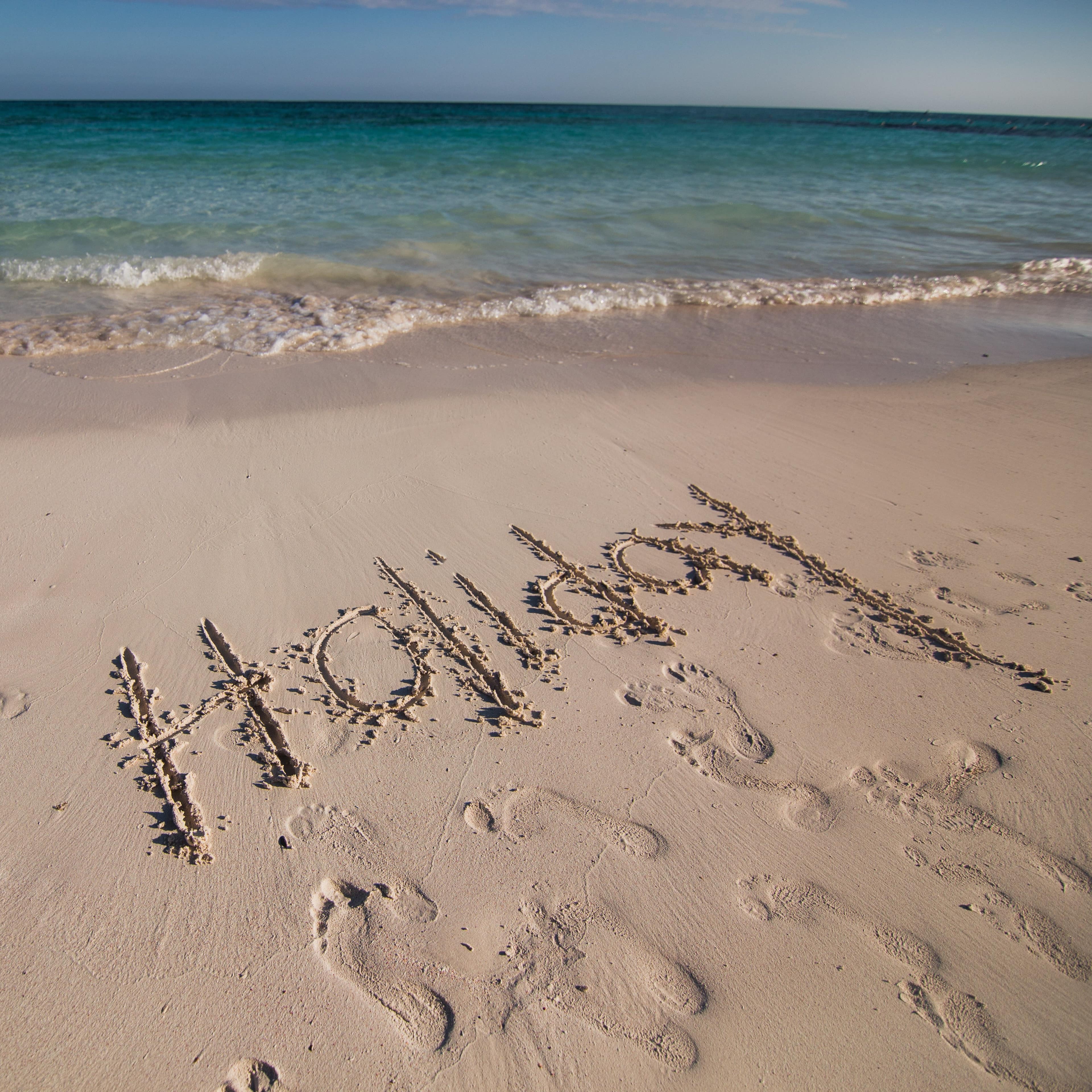 'Holiday' written on soft, pearlescent beach sand with footprints and clear, turquoise water