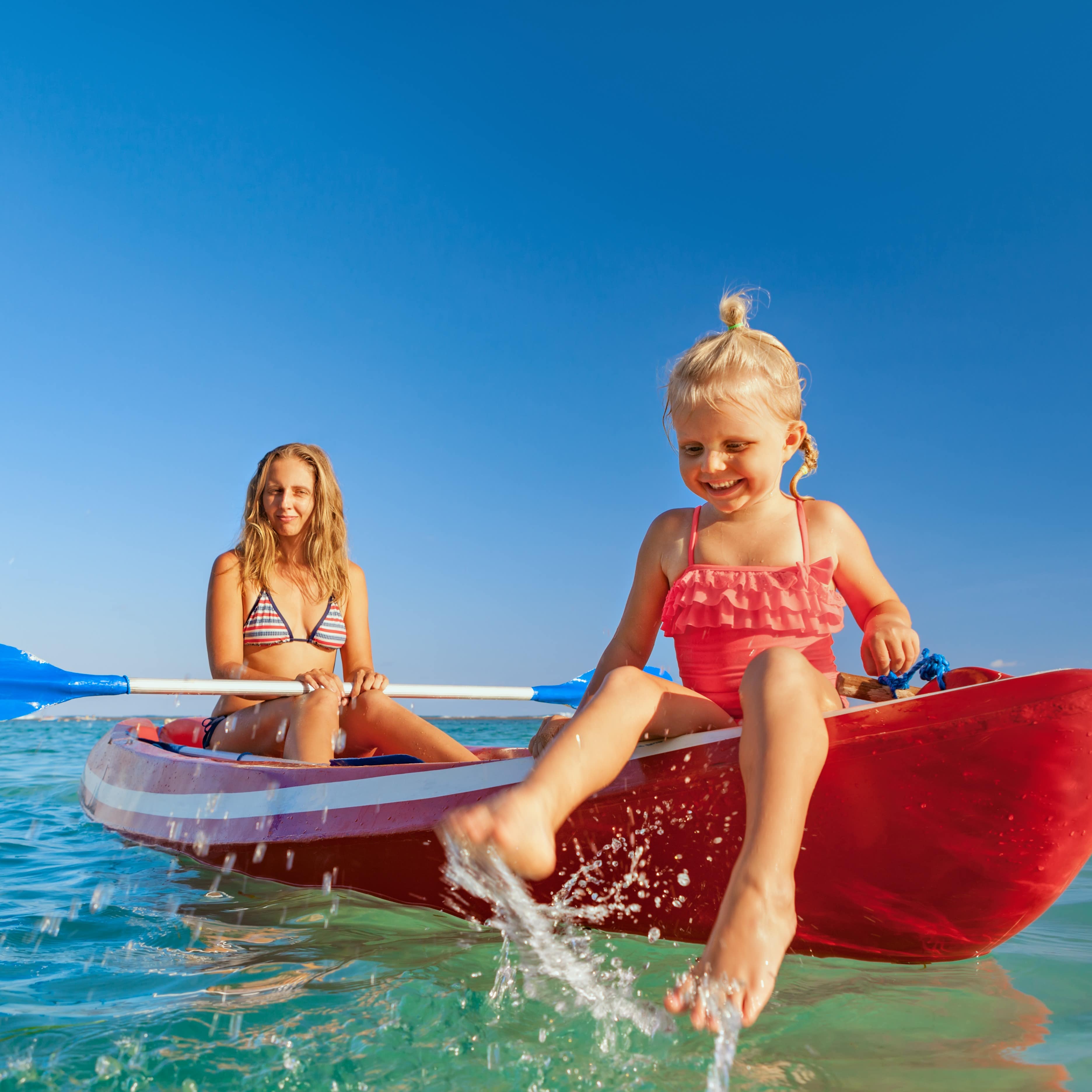Young mother holds paddle and daughter splashes water with feet on kayak in ocean