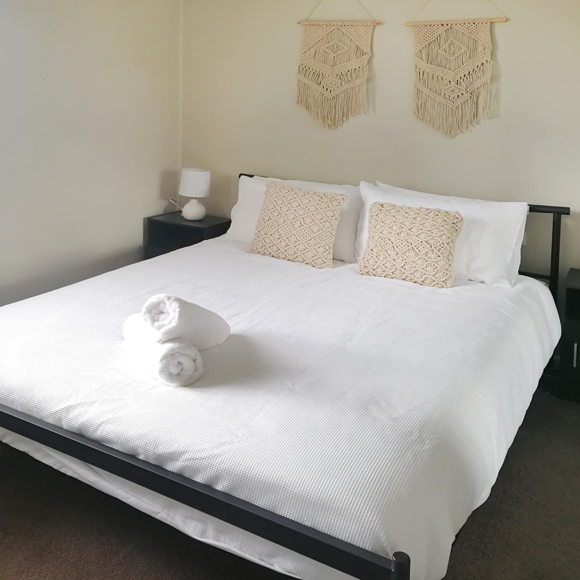 Bedroom with a king size bed, white linen and tan decorative pillows, tan woven wall hangings, neatly rolled towels on bed.