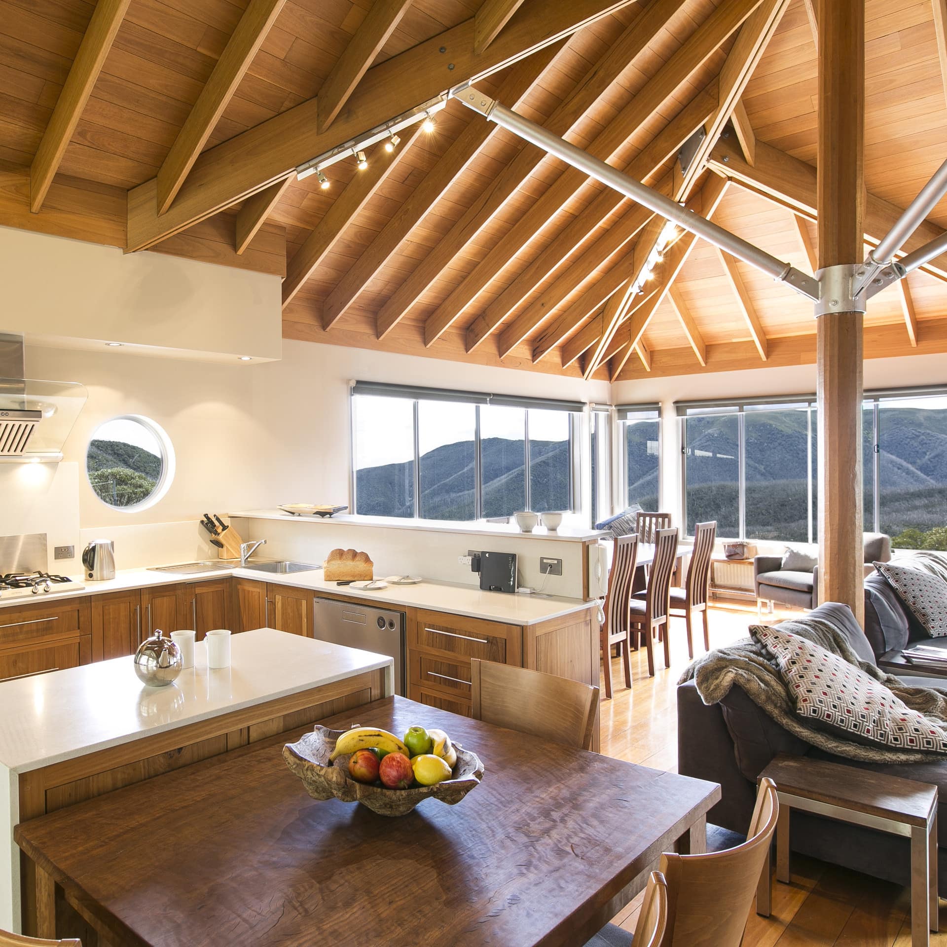 Rustic and modern kitchen and living room with vaulted ceilings, wood furnishings, grey sofas, and panoramic mountain views.