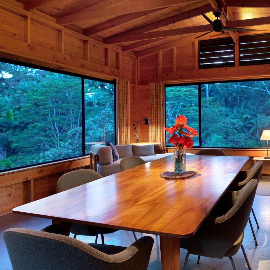 An open dining room with wood panelled walls, wooden dining set, white sofas, and panoramic views of the rainforest.