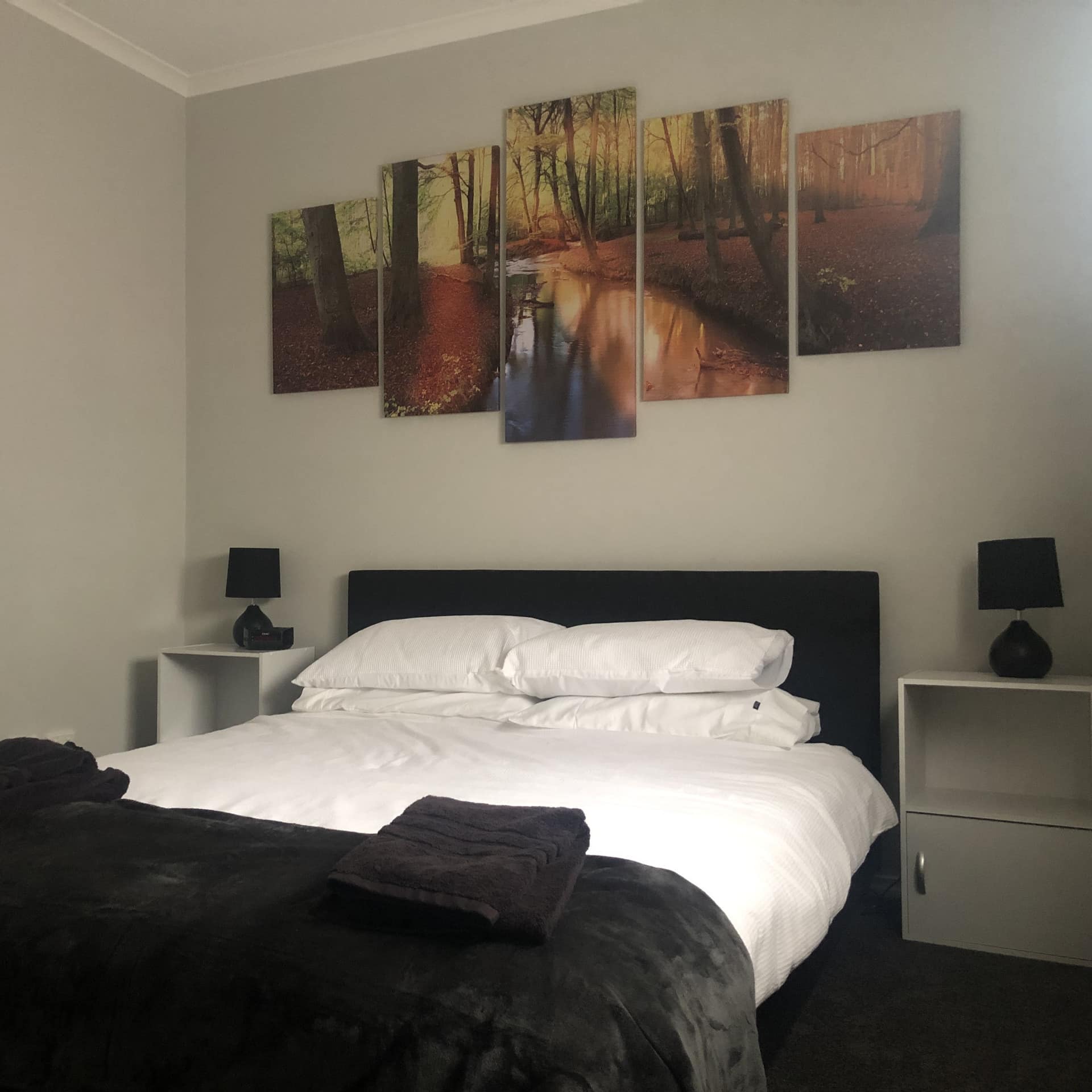 Bedroom with beige walls, a queen-size bed with white and black bedding, and paintings of an autumn forest scene.
