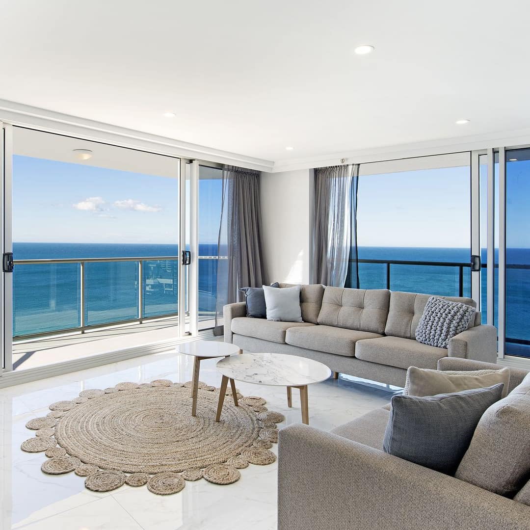 Living room with white stone floors, grey sofas, tan woven rug, and wide windows with panoramic views of the ocean.