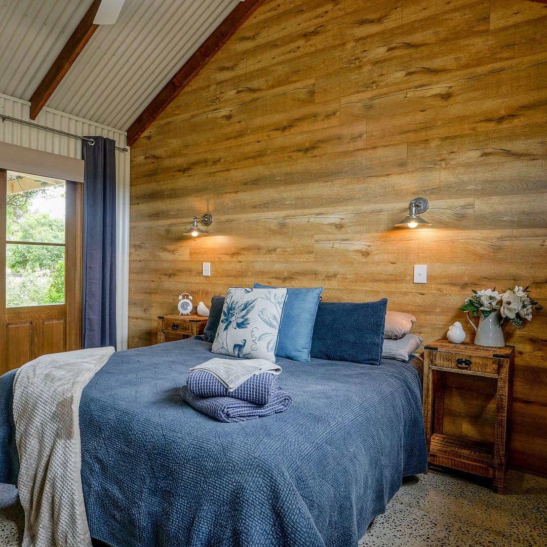 Rustic bedroom with grey carpet, wood-panelled wall, a king-size bed with blue bedding, glass double doors, blue curtains.