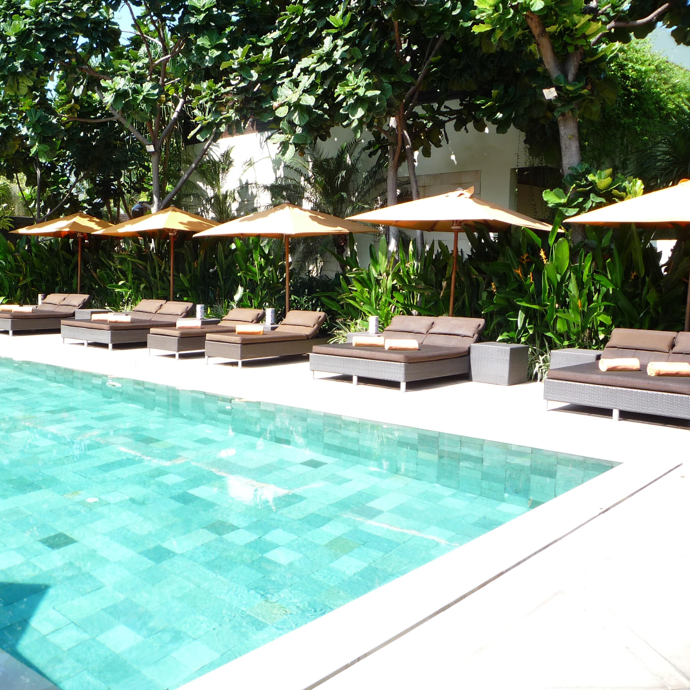 A blue-patchwork-tile pool in front of a white villa with brown lounge chairs, shade umbrellas and tropical plants and trees