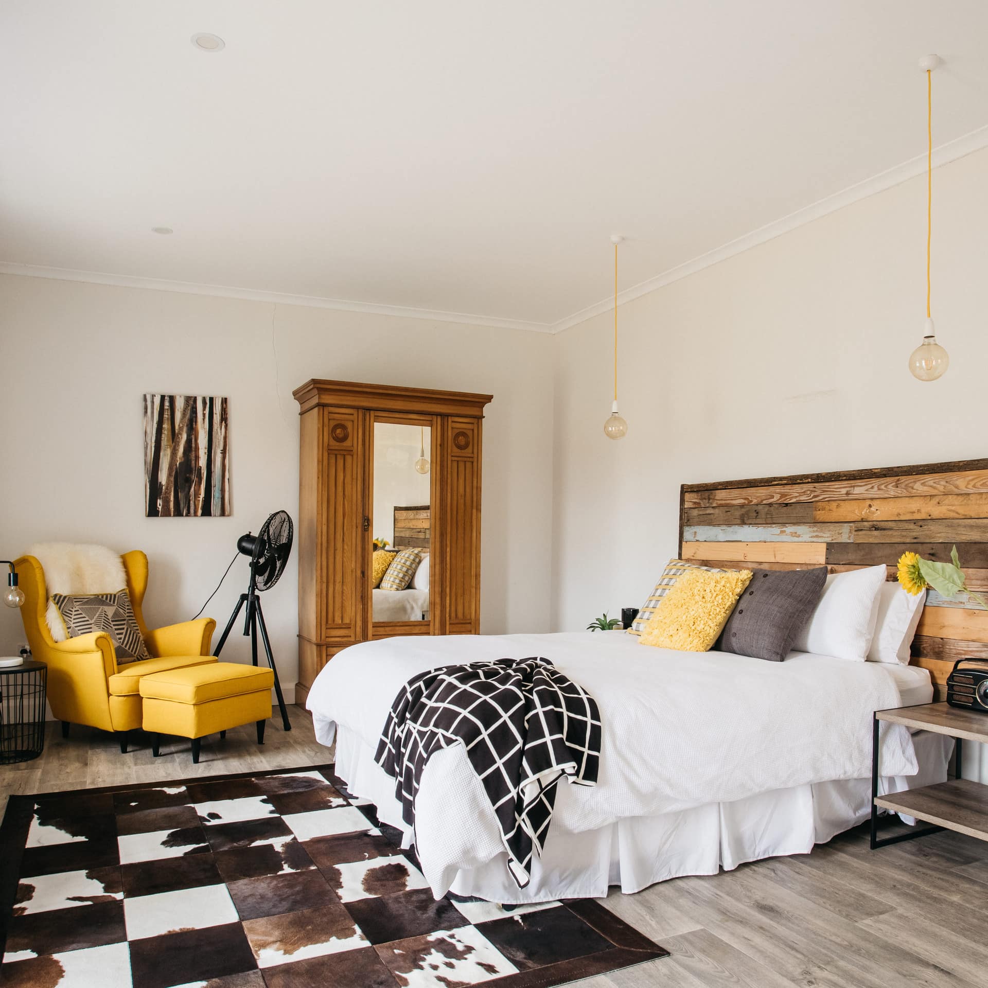 Bedroom with cream walls, a queen size bed with wood panelled headboard, brown and white decor with yellow accents.