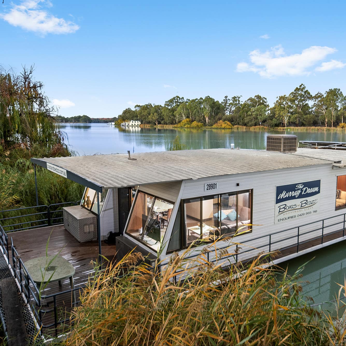 A white houseboat with deck in the Murray River surrounded by trees and lush vegetation.