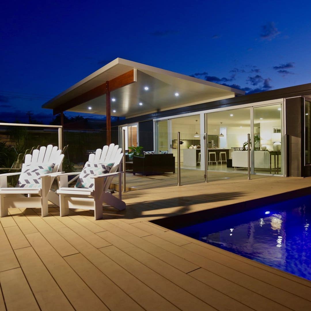 Nighttime view of an inground pool, wooden pool deck with white lounge chairs, and a modern home.