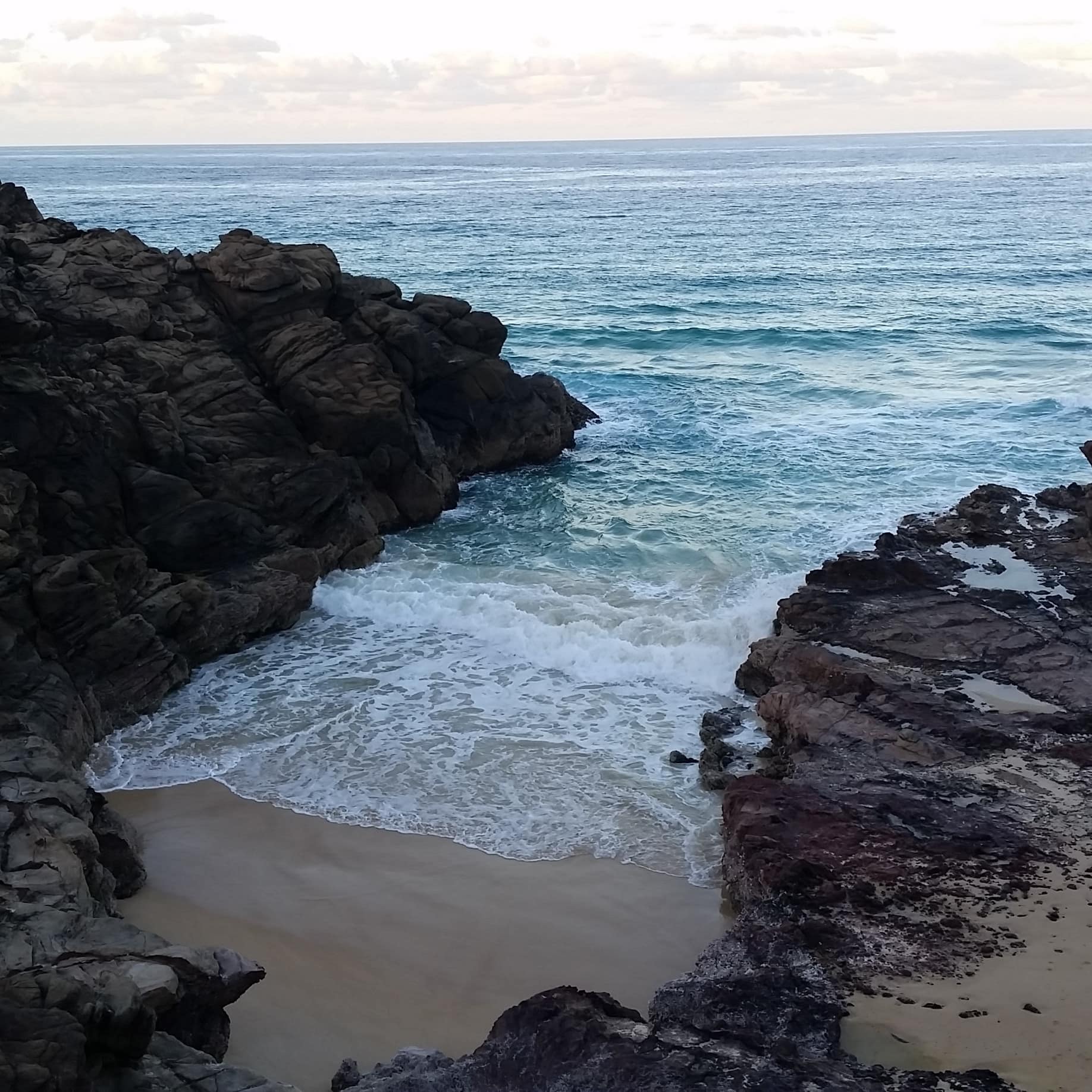 Evening view of a beach with crystal blue water and dark rocky cliffs.