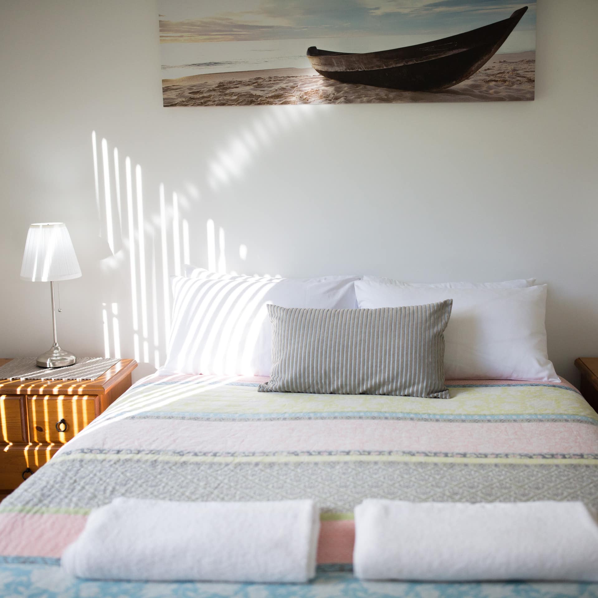 Sunny bedroom with a queen size bed, colorful bedding, wooden bedside tables, and nautical art.