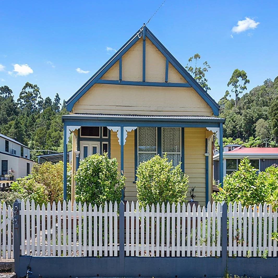 Charming yellow cottage with blue trim, a covered front porch, landscaped lawn, and white picket fence.